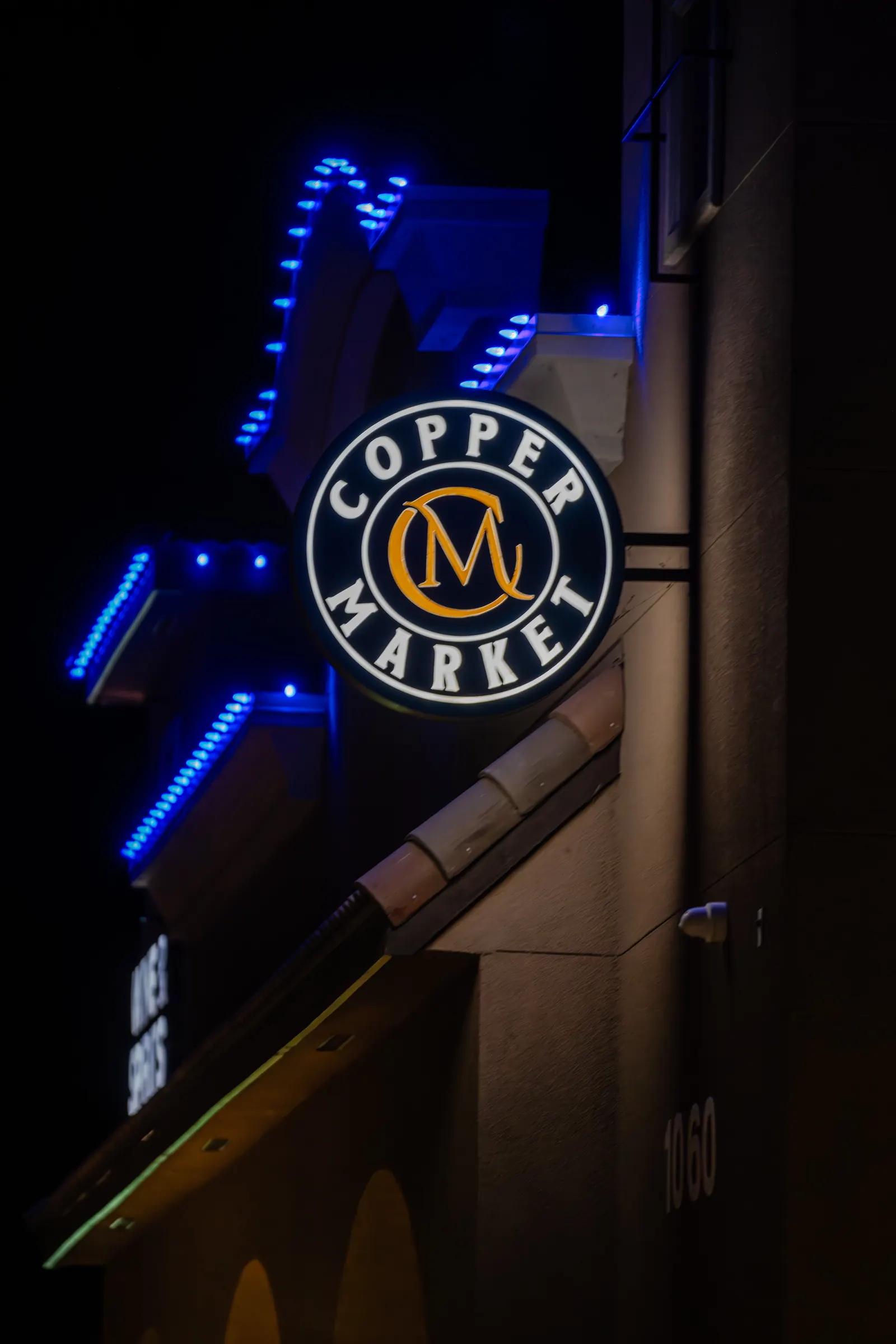 Round Copper Market blade sign lit at night beside blue LED building accents